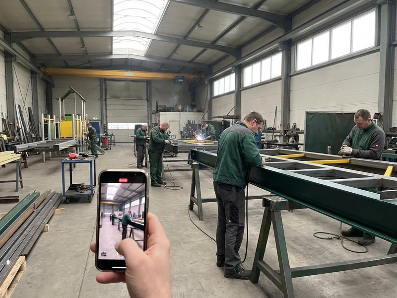 A phone held up in the steel workshop streaming a live video tour of welders at a frame