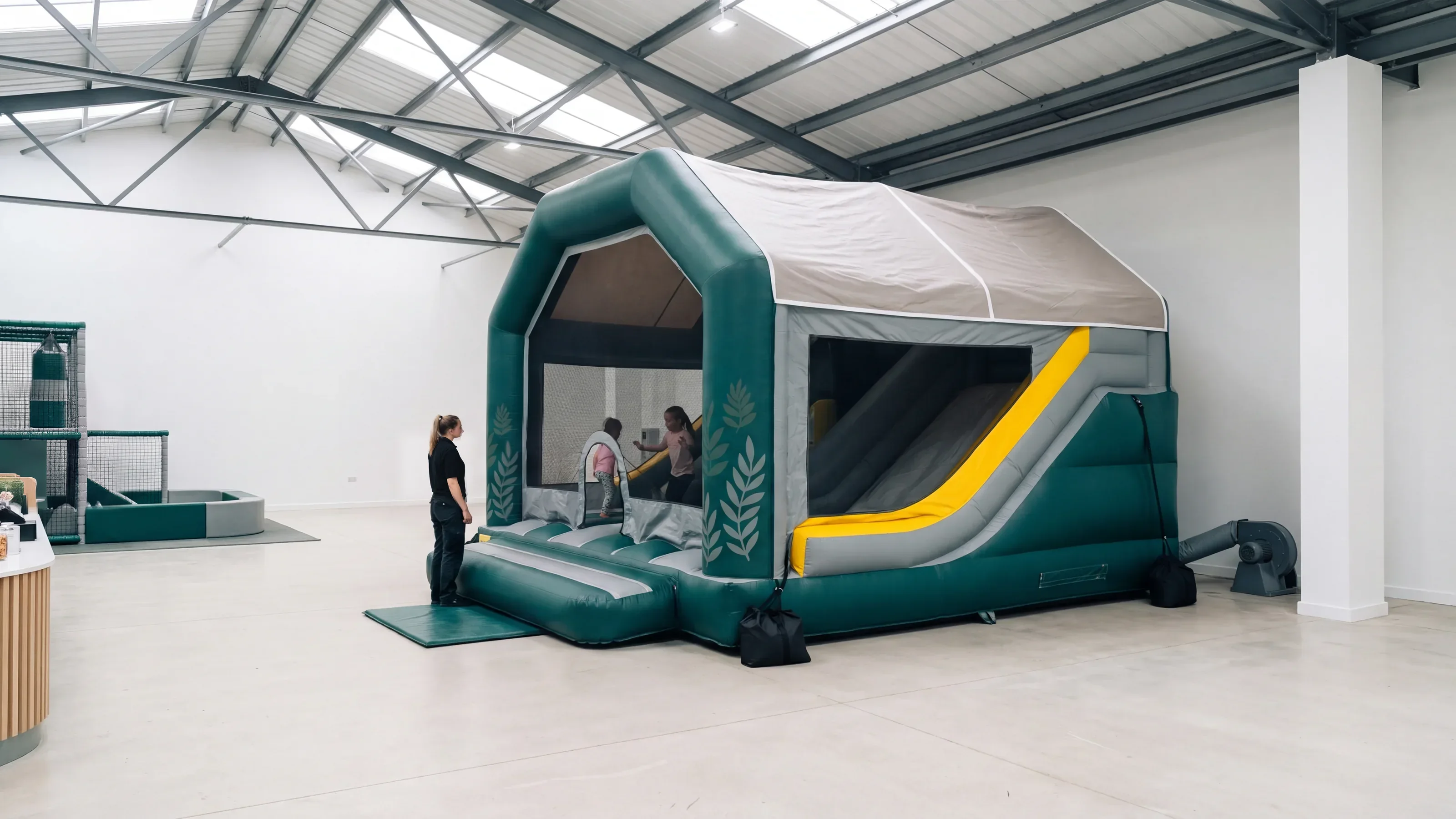 A commercial combo inflatable in deep green and grey inside a skylit family entertainment center, its blower connected at the back