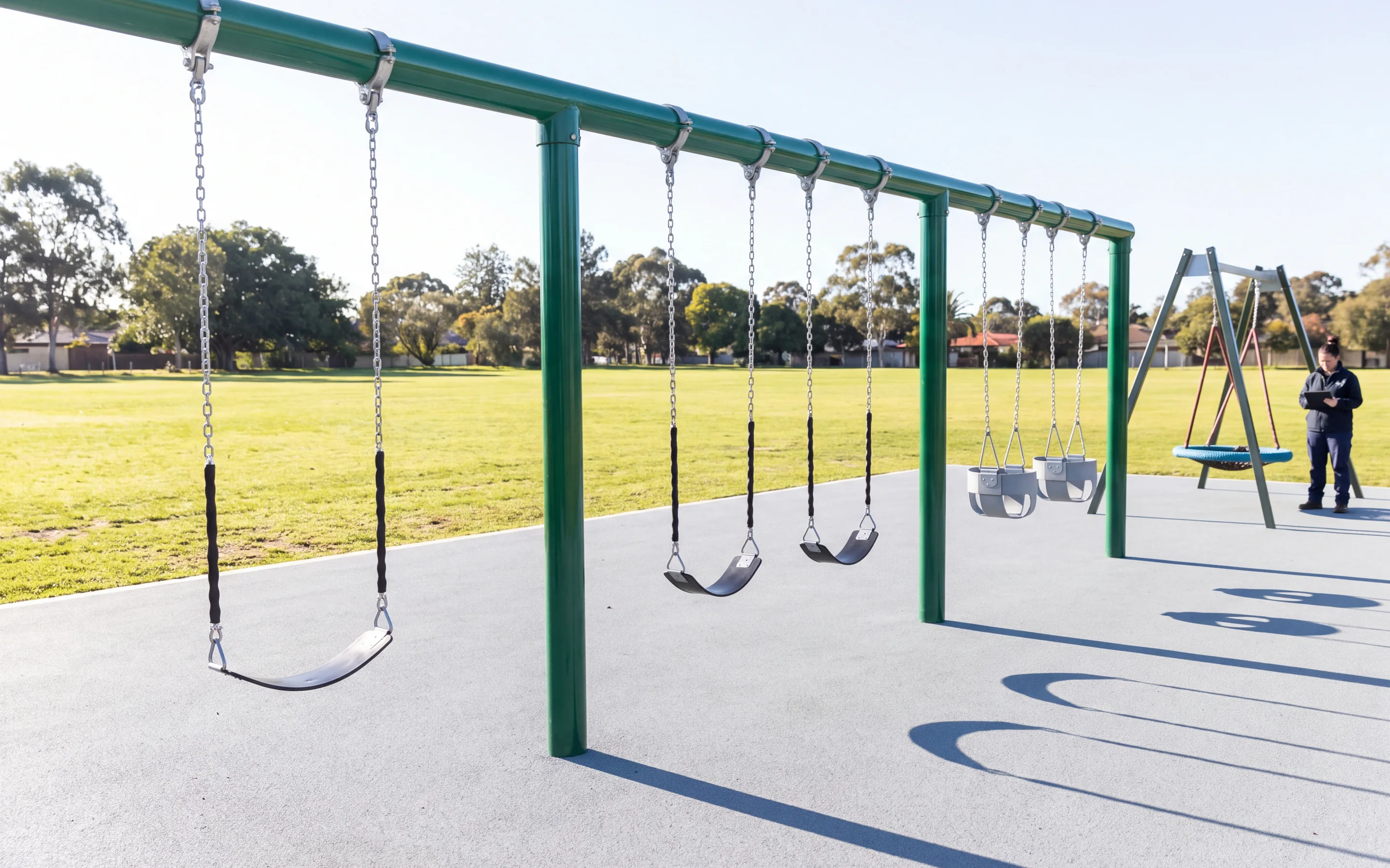 A three-bay evergreen straight-post swing frame in a morning park, two black belt seats in one bay and two grey toddler bucket seats in the next, with a separate nest swing on its own frame behind