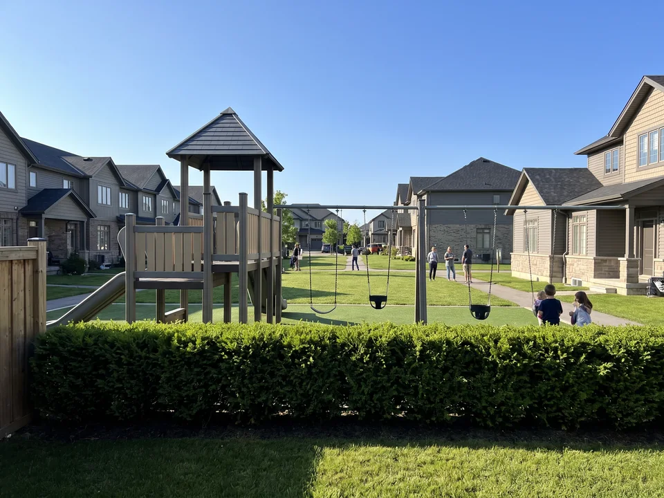 View from behind a low hedge in a back yard across the green to a play structure whose tower faces the path