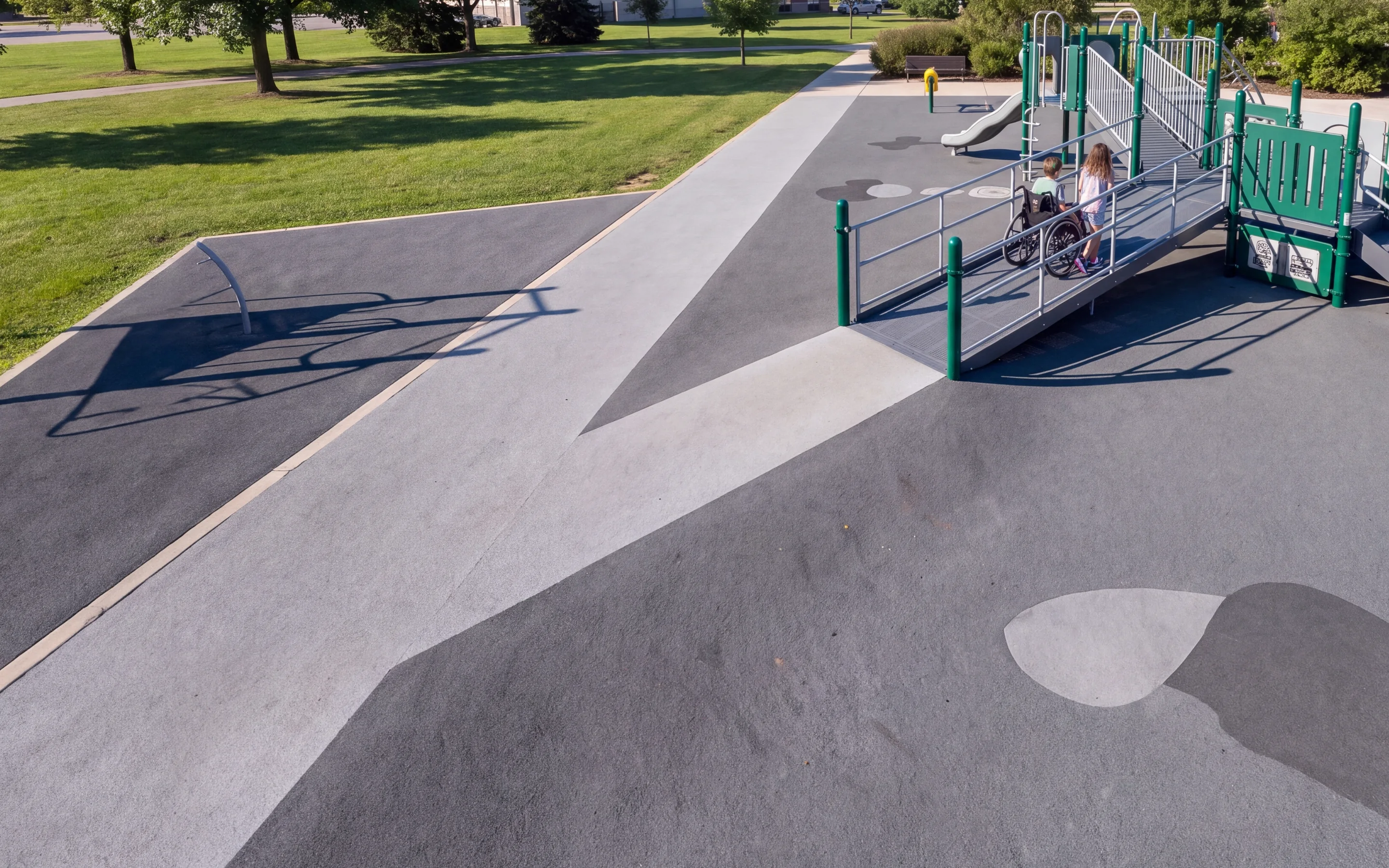 A child using a wheelchair and a friend walking side by side up a solid anti-slip ramp to an evergreen play structure, with a transfer platform beyond