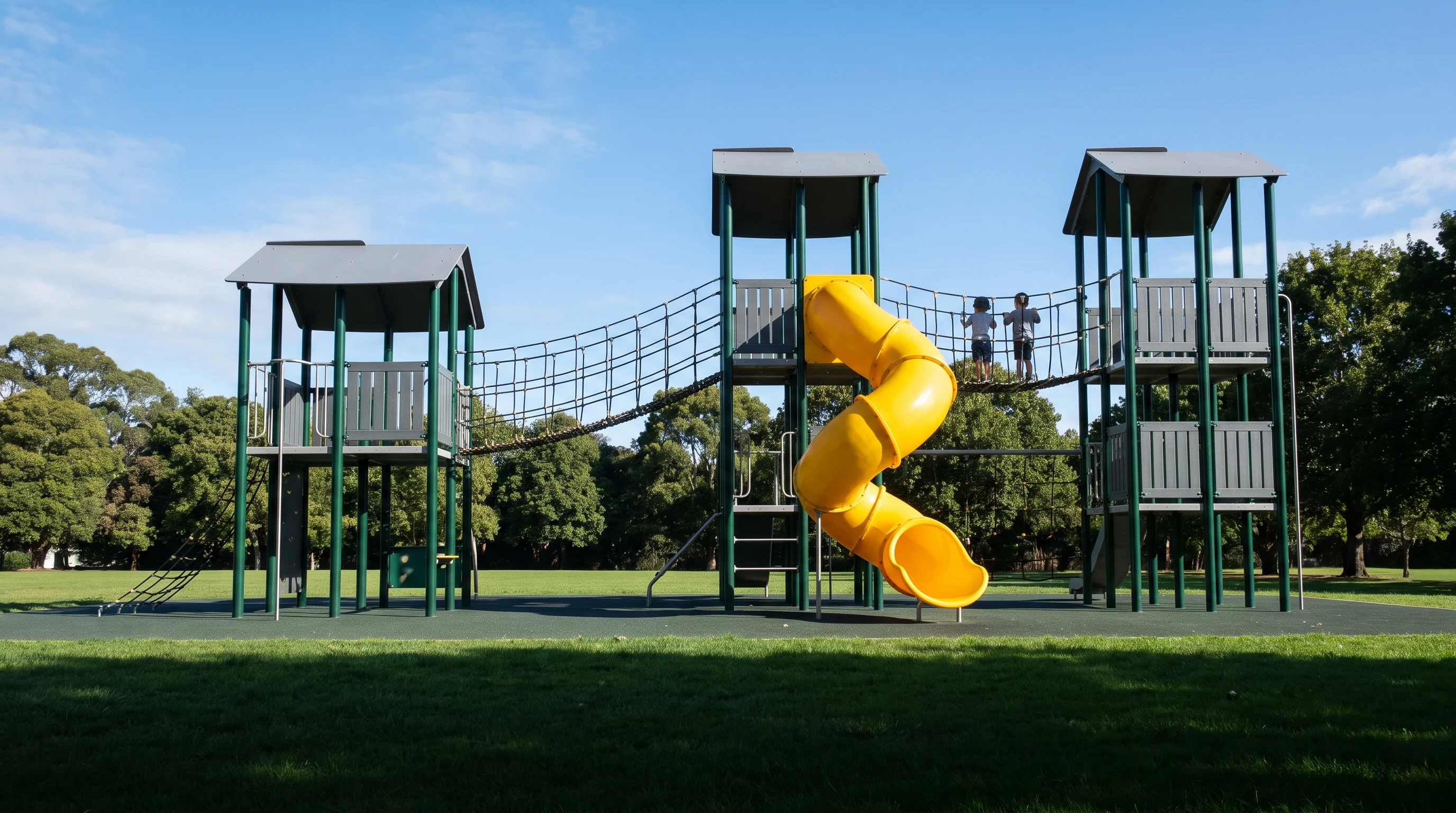 Large commercial multi-deck playground structure with evergreen posts, grey HDPE panels and a yellow tube slide in a public park