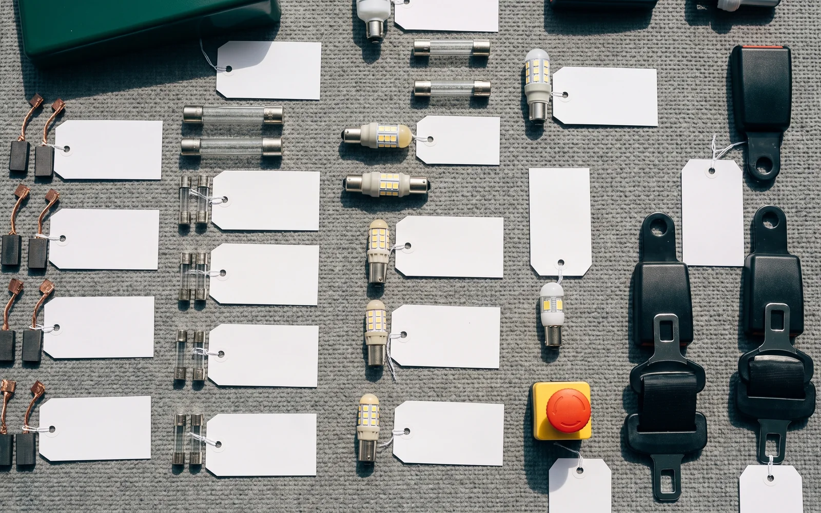 A kiddie ride wear-parts set laid out on a grey mat: carbon brushes, fuses, LED bulbs, seat-belt buckles and a spare emergency-stop button, each with a blank tag