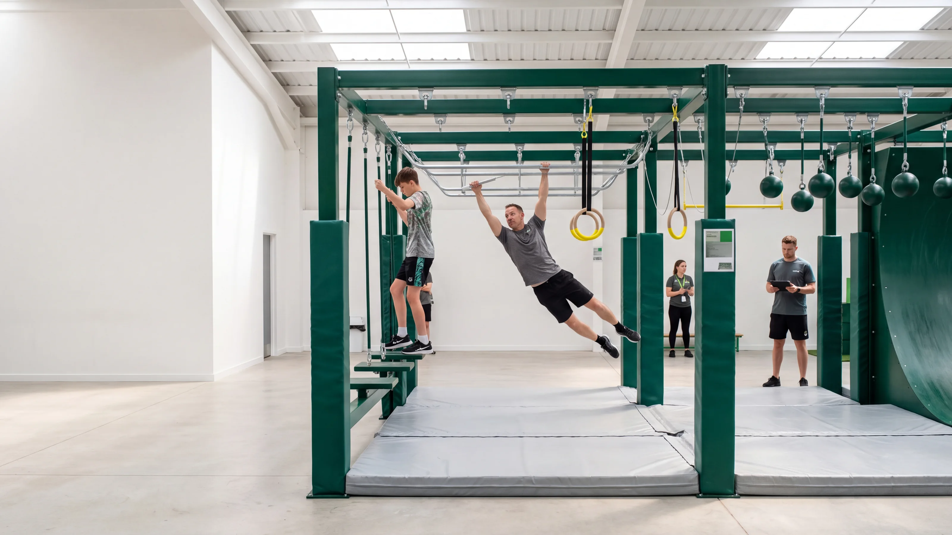 A skylit ninja gym with a long evergreen steel rig: an adult swinging on a lache bar, a teenager on floating steps, a pair of rings with yellow grips hanging from one hang point