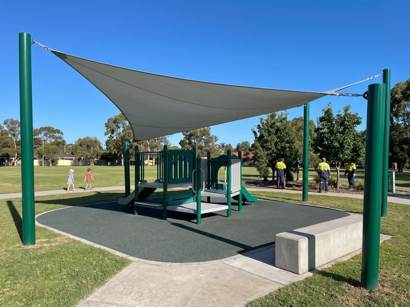 A shade sail on four evergreen posts set outside the surfacing, its shadow covering the play structure decks