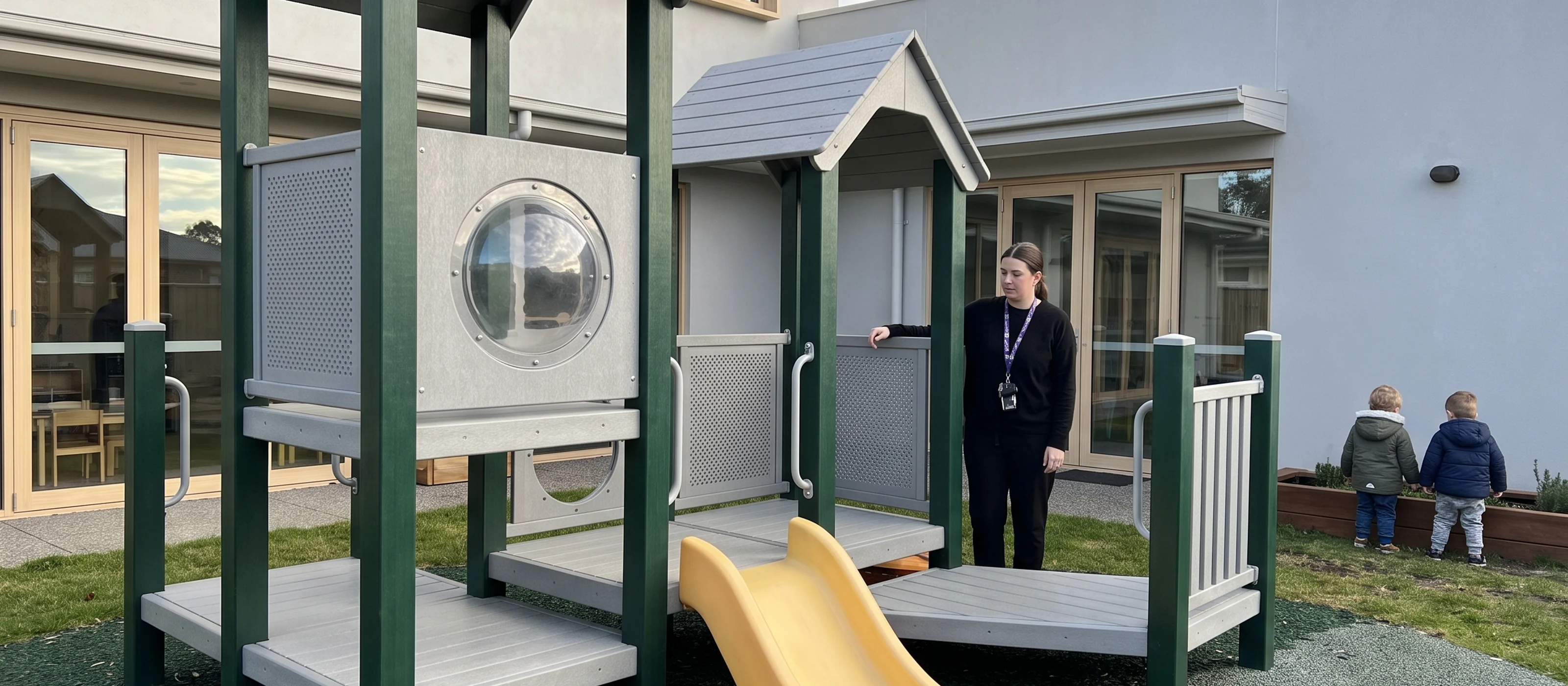 A child care center yard seen from a child's eye level: a sand and water table, a low-deck preschool structure and a teacher with her hand on a deck guardrail