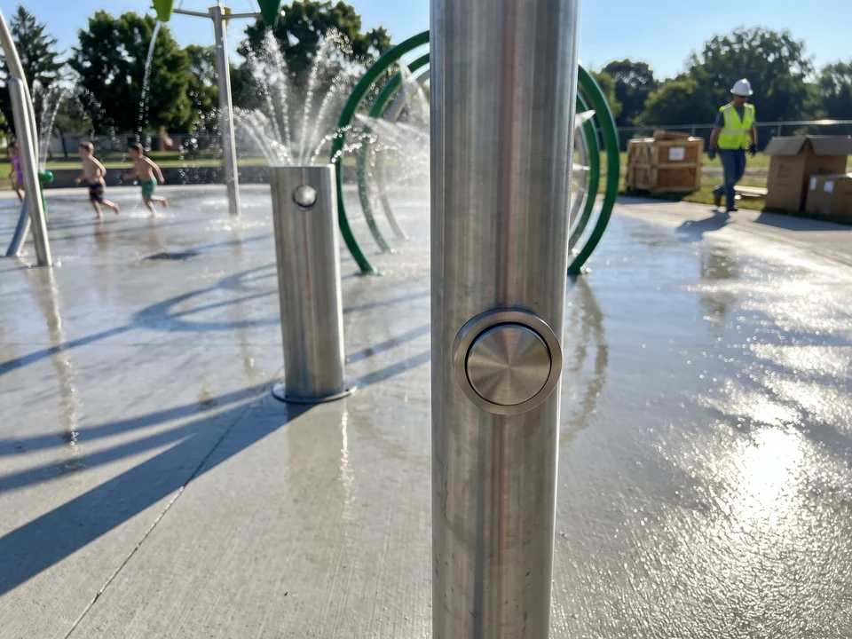 Close view of a stainless activation bollard with its push button at the edge of a splash pad