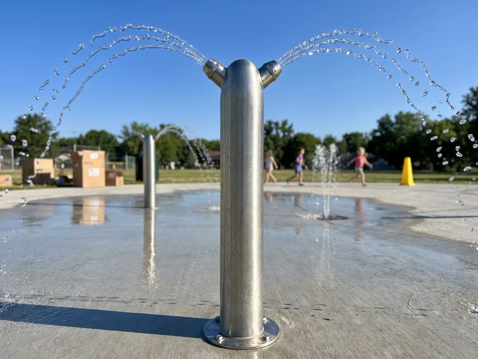 A brushed 304 stainless spray post on its base plate throwing water arcs from two nozzles at the top