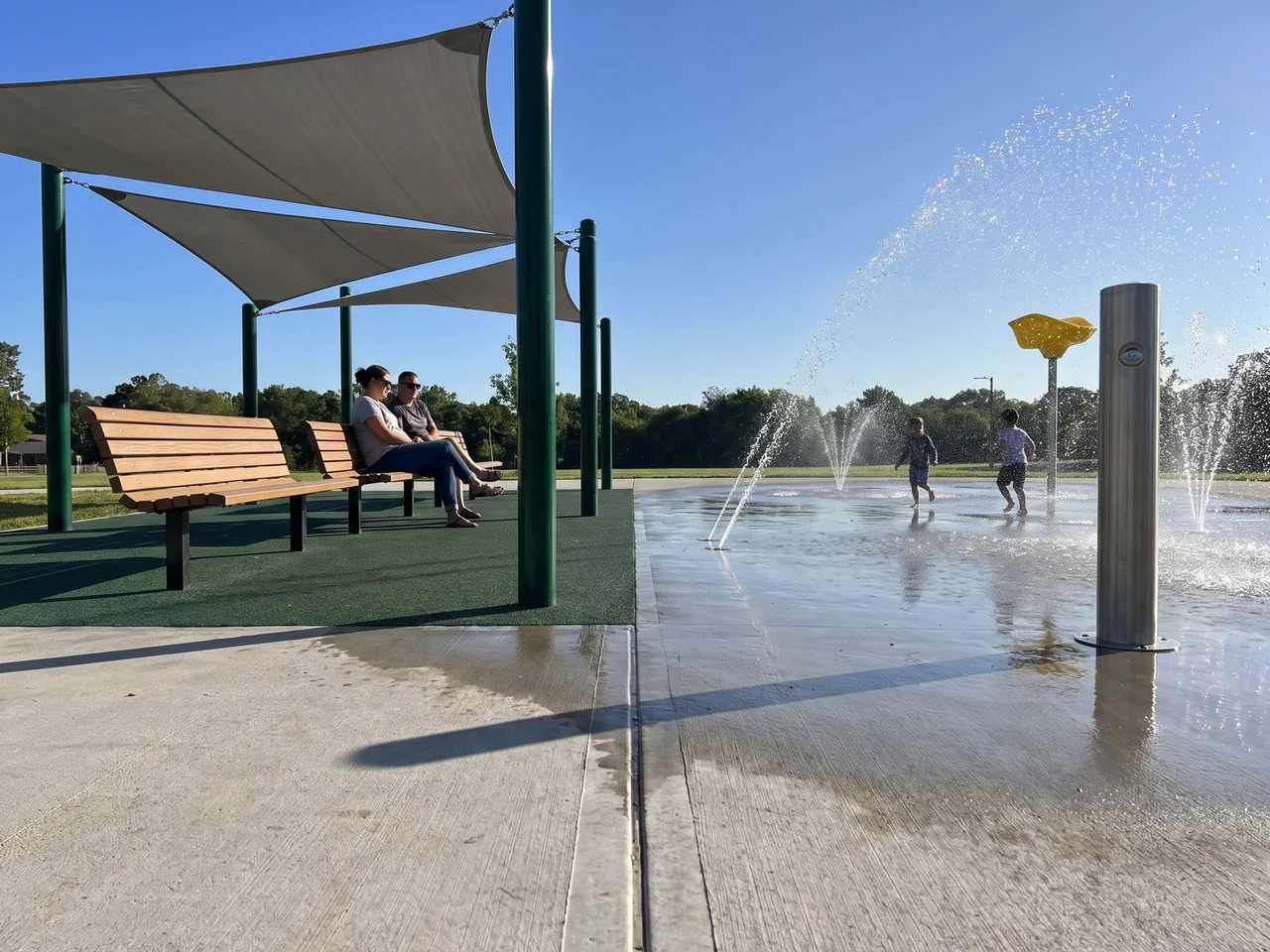 The dry edge of a splash pad: benches under a shade sail beside the wet pad, with rubber play surfacing beyond