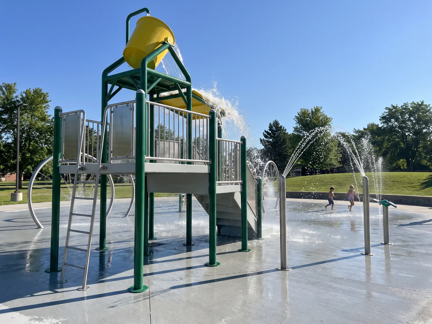 A multi-feature water play structure with an evergreen fiberglass frame, light grey platforms, a ladder and a large tipping bucket, two children running on the pad beyond