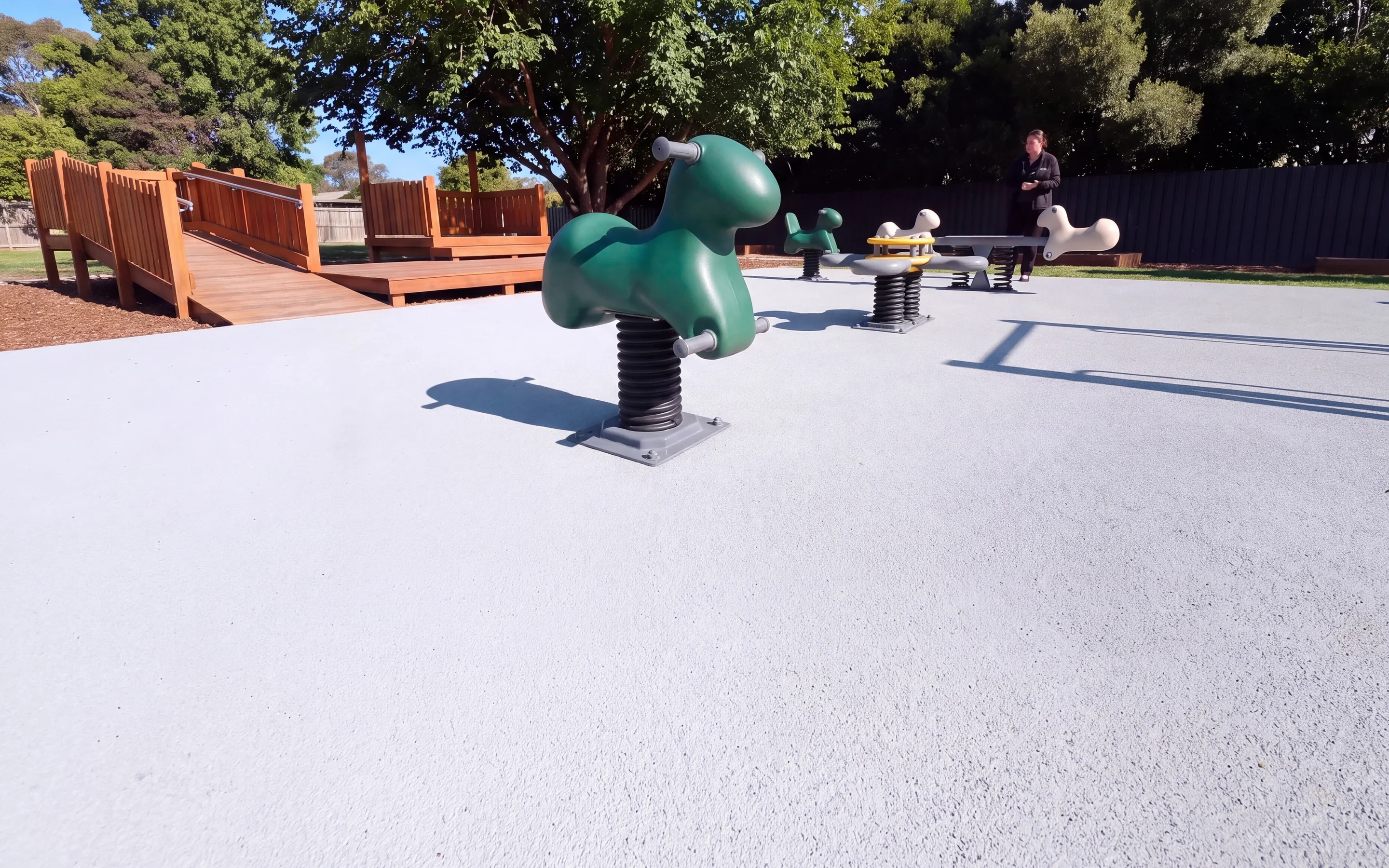 A single-seat evergreen spring rider on a black rubber pinch-guard sleeve, its base plate on light grey poured rubber surfacing in a morning park, with a second rider, a four-seat platform rider and a seesaw behind