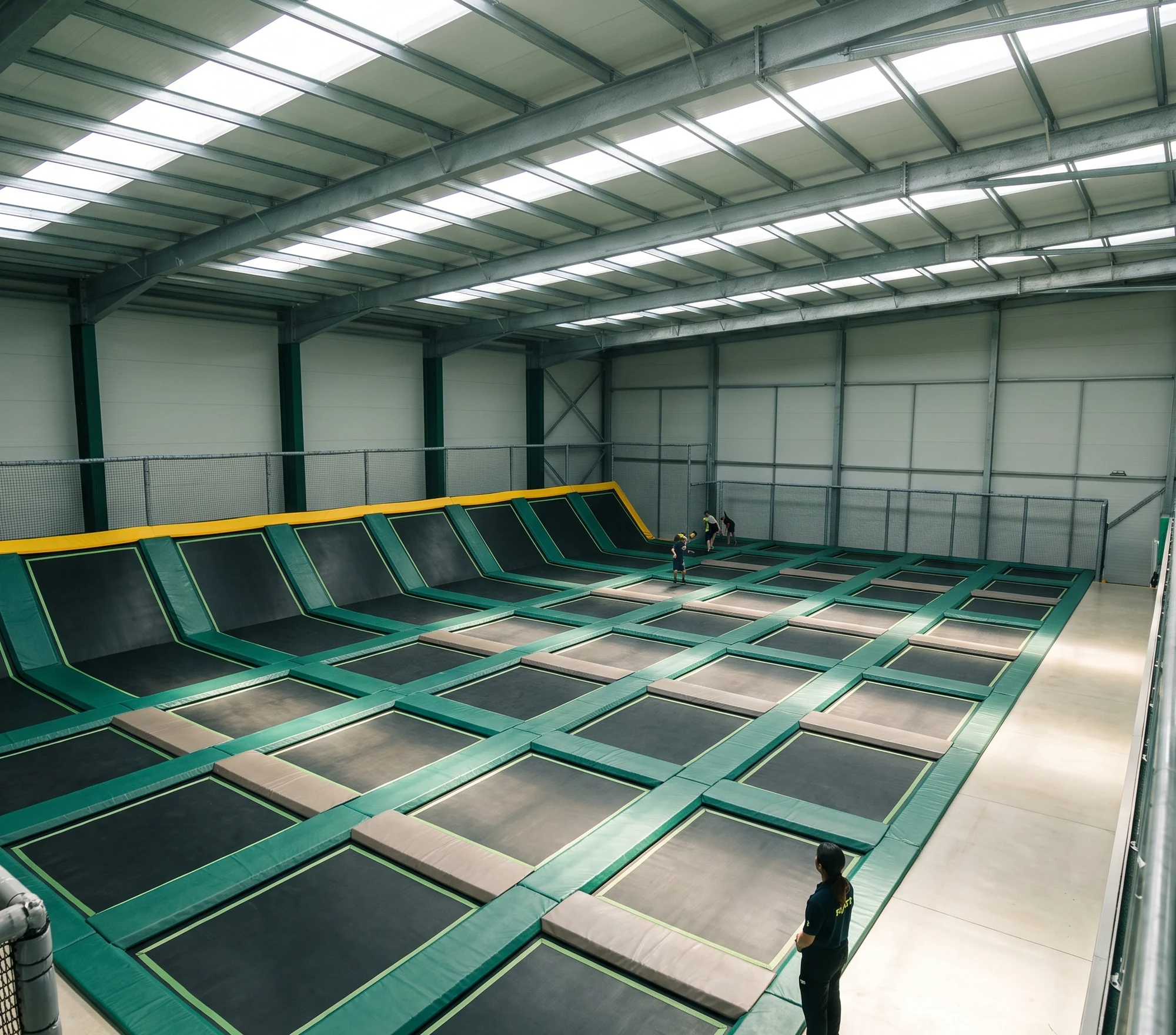 Main trampoline court seen from a mezzanine: black jump beds in a grid of evergreen frame pads, angled wall beds on the far wall, a foam pit and a netted dodgeball court